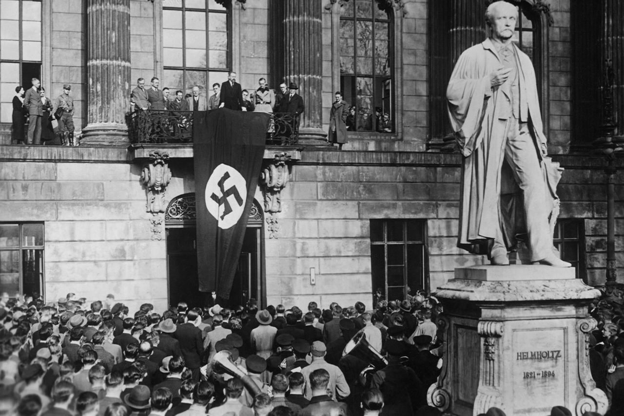 A photograph of the inner courtyard of Friedrich Wilhelm University in Berlin. There are many people in the courtyard. Eugen Fischer is standing on the balcony, giving a speech. A large banner with a swastika is hanging from the parapet.