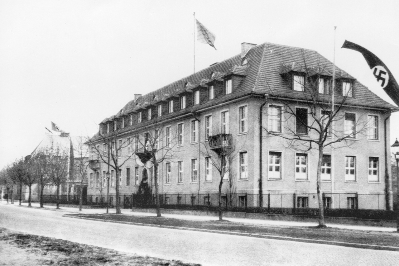 Schwarz-Weiß-Foto. Perspektive von der Ihnestraße auf das Hauptgebäude des Kaiser-Wilhelm-Instituts für Anthropologie, menschliche Erblehre und Eugenik um 1933/34. Auf einer Fahnenstange weht die Hakenkreuzflagge.