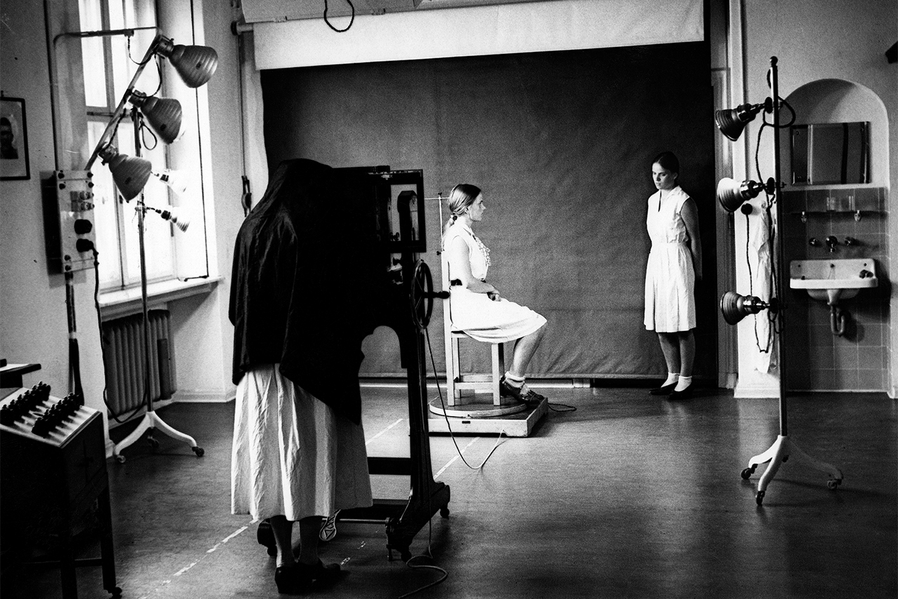 Photograph of three individuals in an examination room. One person wearing a dress or a long white lab coat is taking a picture from the side of a person sitting on a chair. The twin sister of the person being photographed is standing to one side.