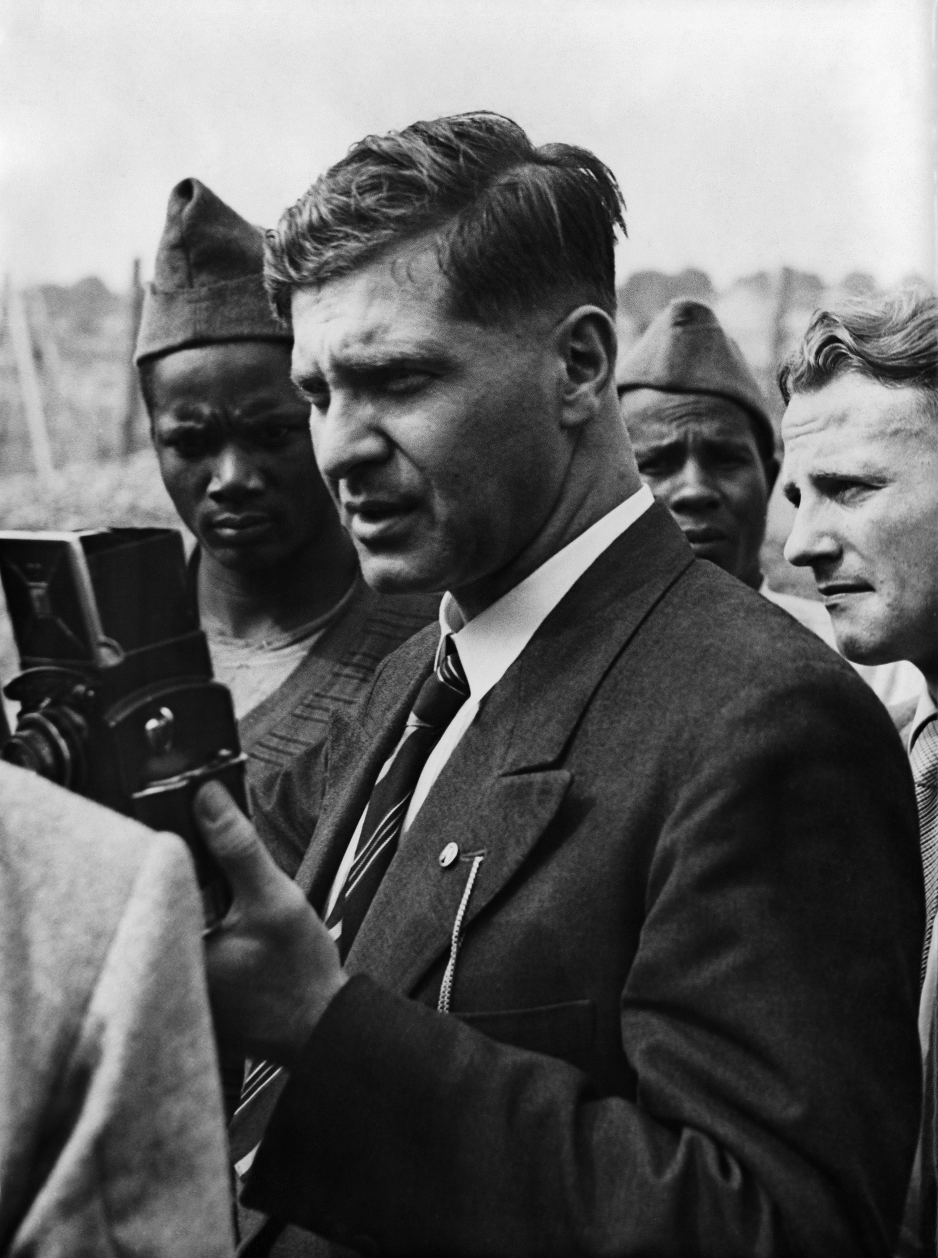 A black-and-white photograph of a small group of people. Wolfgang Abel is at the front. He is a White man wearing a black suit and necktie. Two people with dark skin wearing prison clothing are standing behind him.