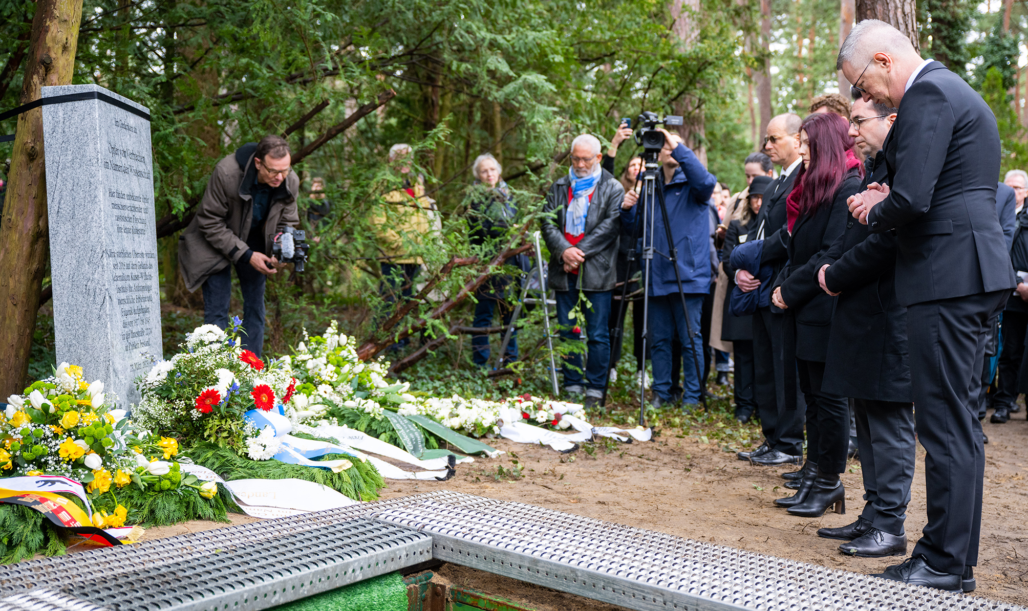 The photograph shows people at a cemetery. They include the president of Freie Universität, Günter Ziegler. The group is standing in front of a memorial stone surrounded by wreaths. Many people have their hands folded. Two people are taking pictures.