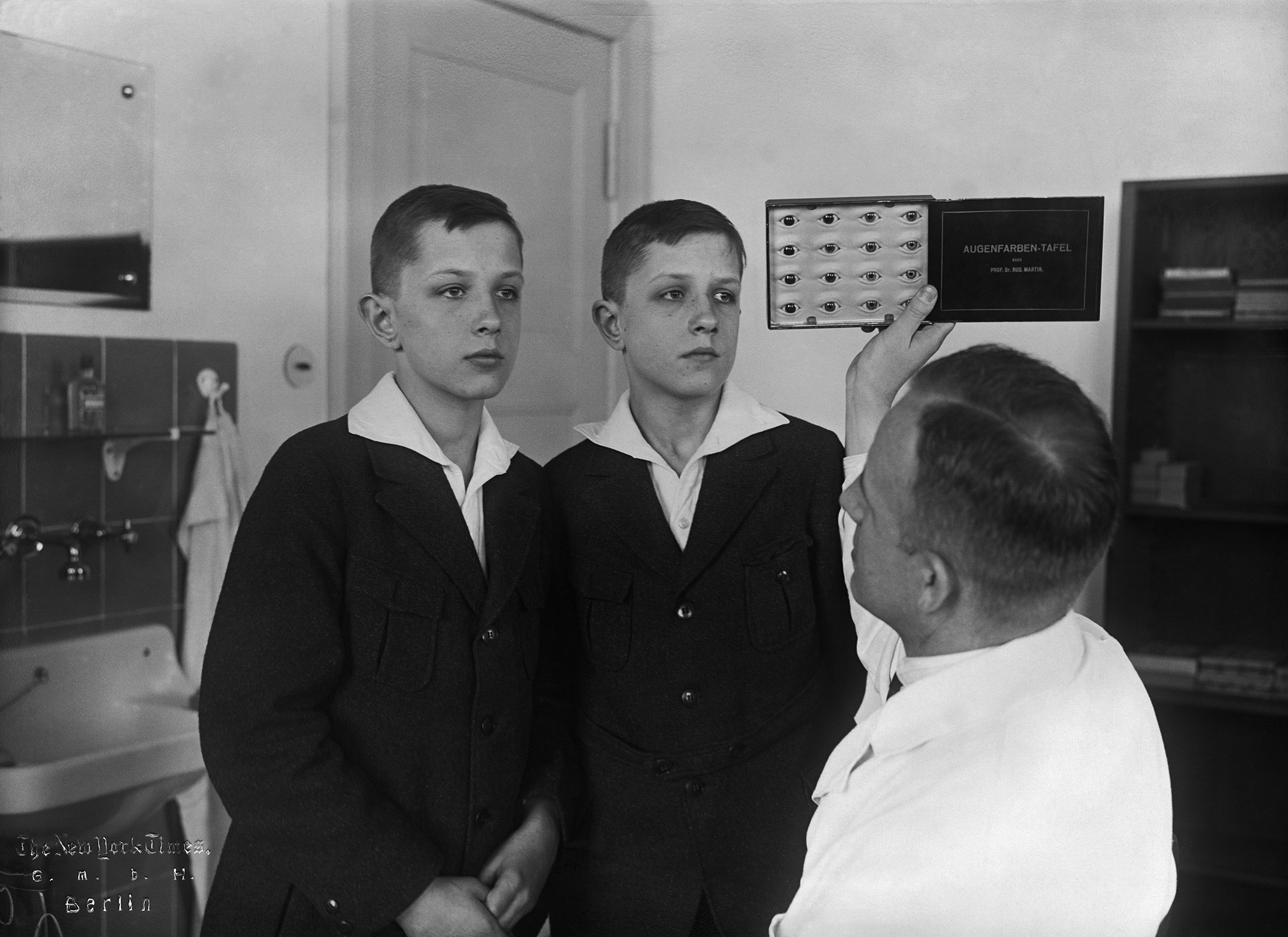 Foto von Otmar von Verschuer und einem minderjährigen Zwillingspaar. Die Zwillinge sehen sich sehr ähnlich und sind gleich angezogen. Verschuer hält eine Tafel mit Nahaufnahmen von sechzehn Augen, die er mit den Augen der Zwillinge abgleicht.