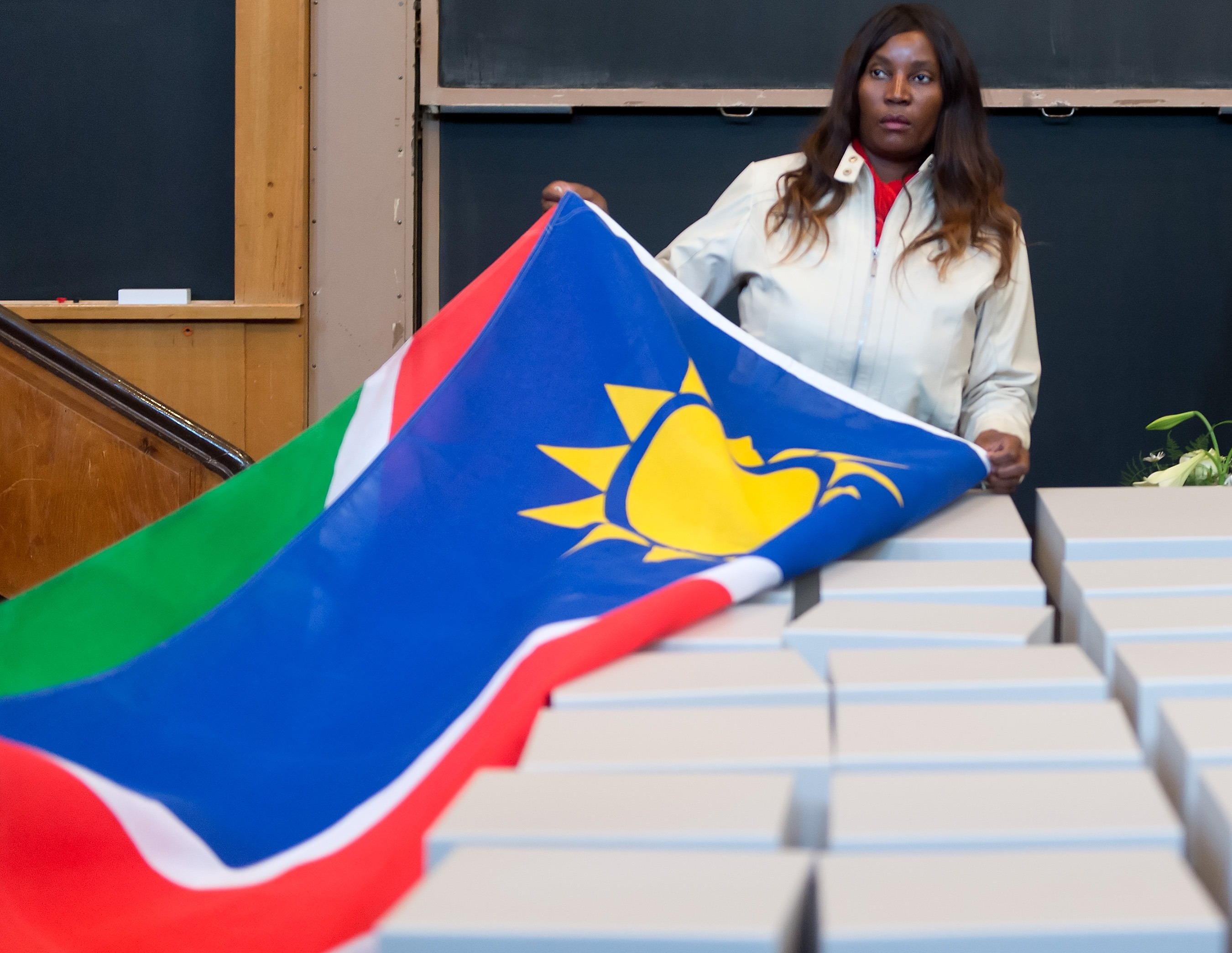 Foto einer Person mit dunkler Haut, welche die Flagge Namibias von einigen weißen Kisten zieht, die vor ihr aufgereiht stehen.