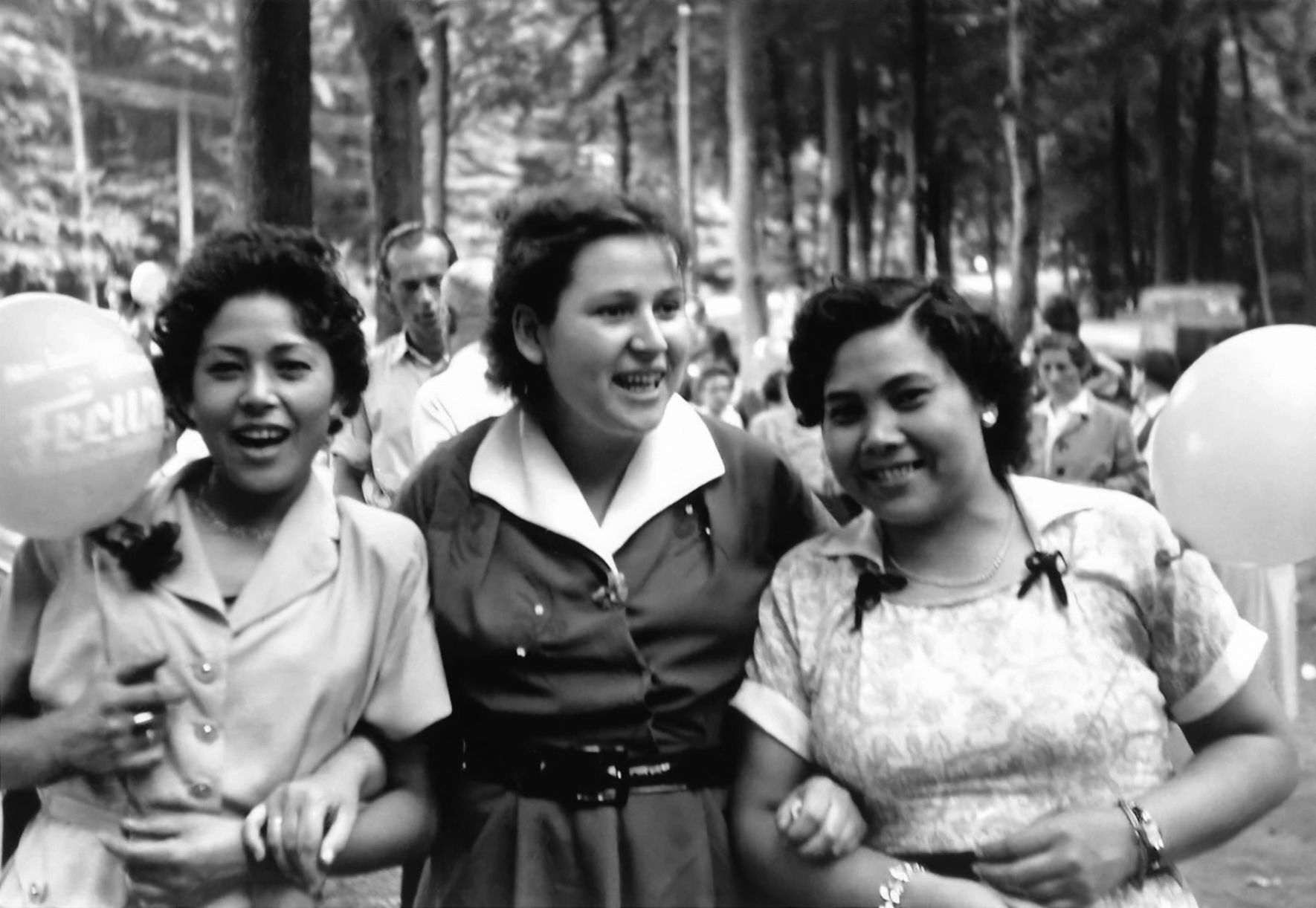 A black-and-white photograph of the sisters Cäcilie, Elfriede, and Luzie Borinski taken in 1955. Elfriede Borinski has light-colored skin, dark hair and is wearing a dark dress. Cäcilie and Luzie Borinski have darker skin and are wearing light-colored clothing. They are smiling toward the camera and holding balloons in their hands.