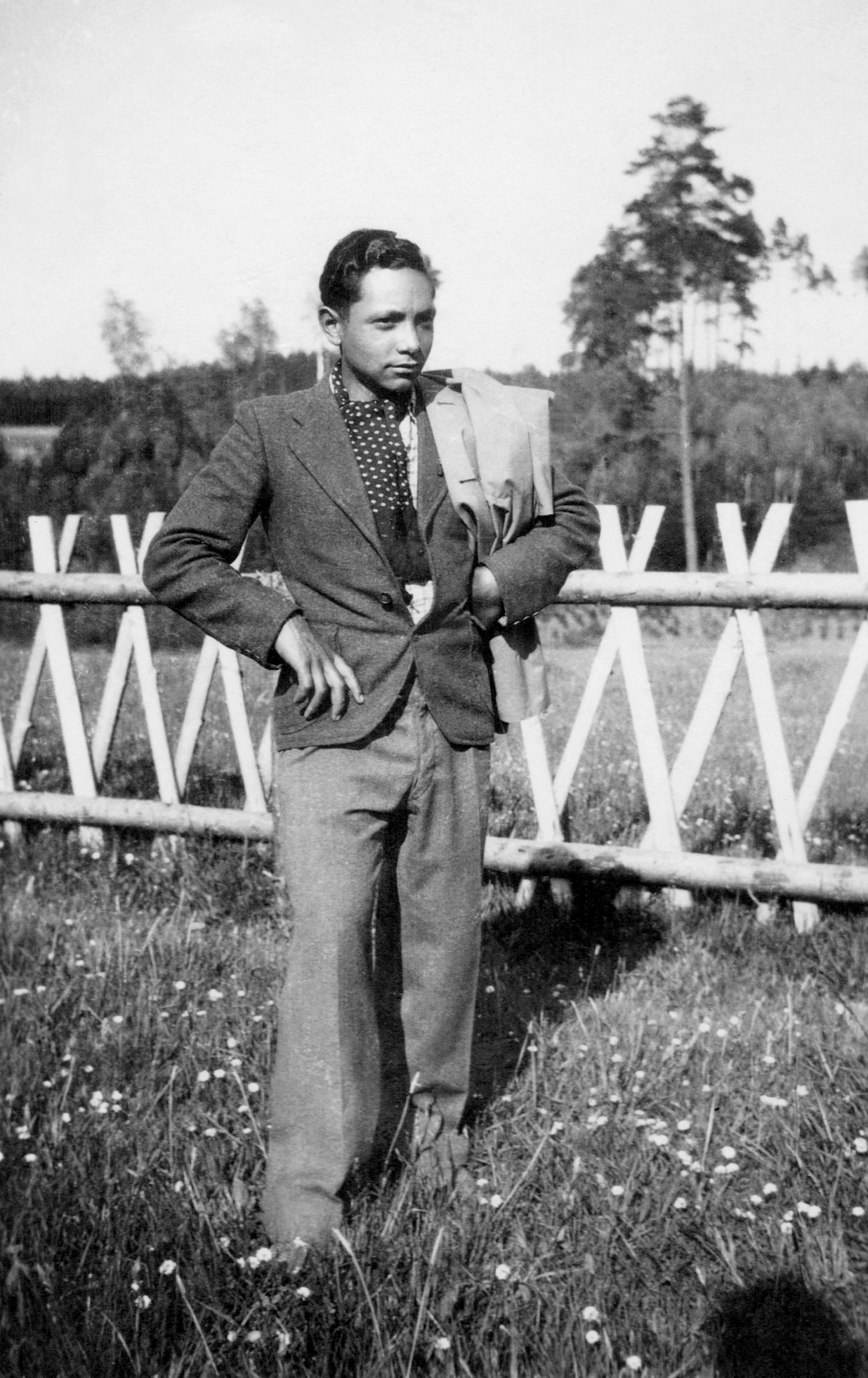 This image is a black-and-white photograph of a young man with dark skin. He is wearing a suit and standing in a meadow in front of a fence.