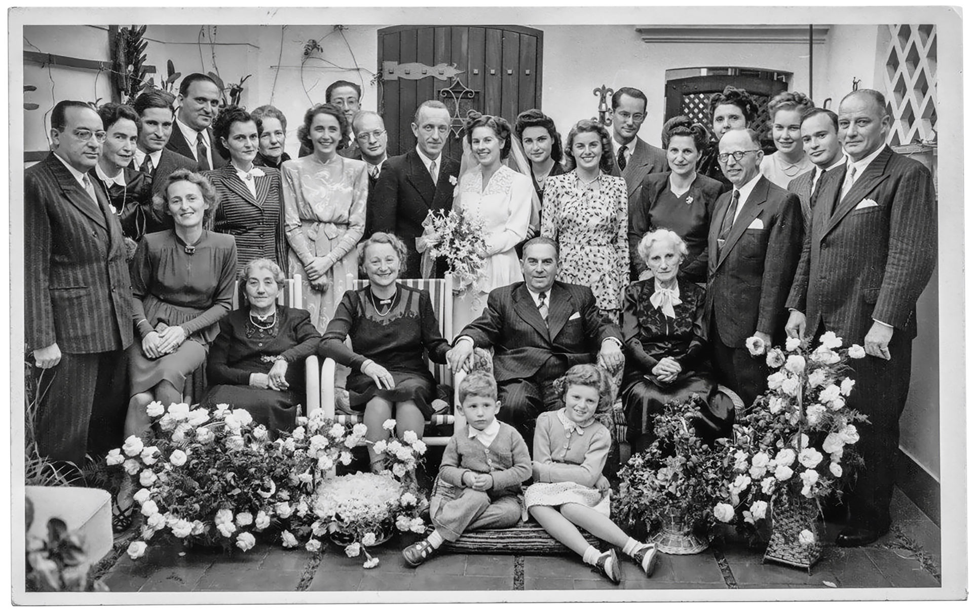 Foto einer großen Menschengruppe, die festlich gekleidet ist. In der Mitte steht ein Brautpaar, umgeben von einigen großen Blumensträußen.