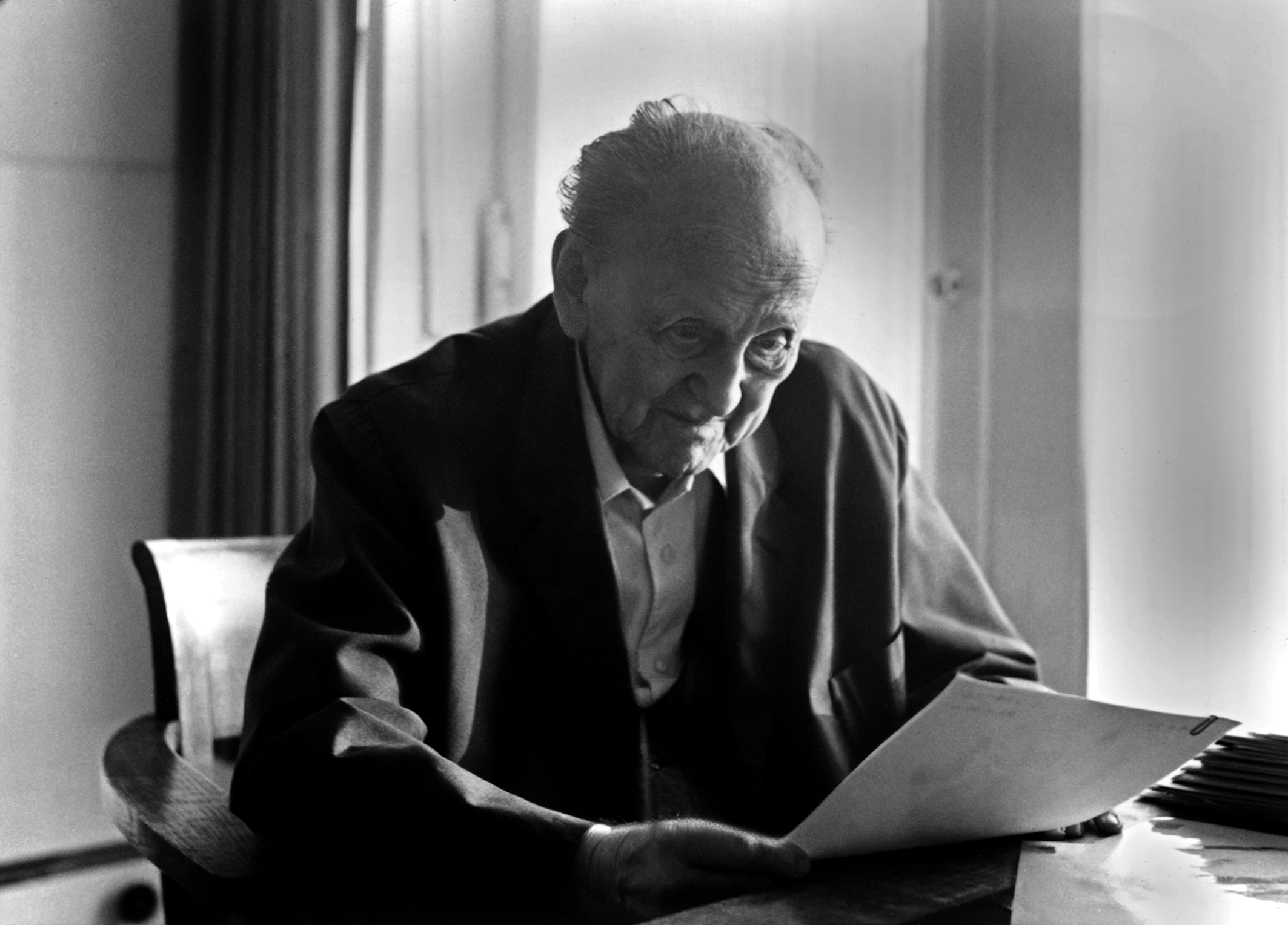 This photograph shows an older White man sitting at a desk. He is wearing a dark jacket and a white shirt and is looking as a sheet of paper in his hands.