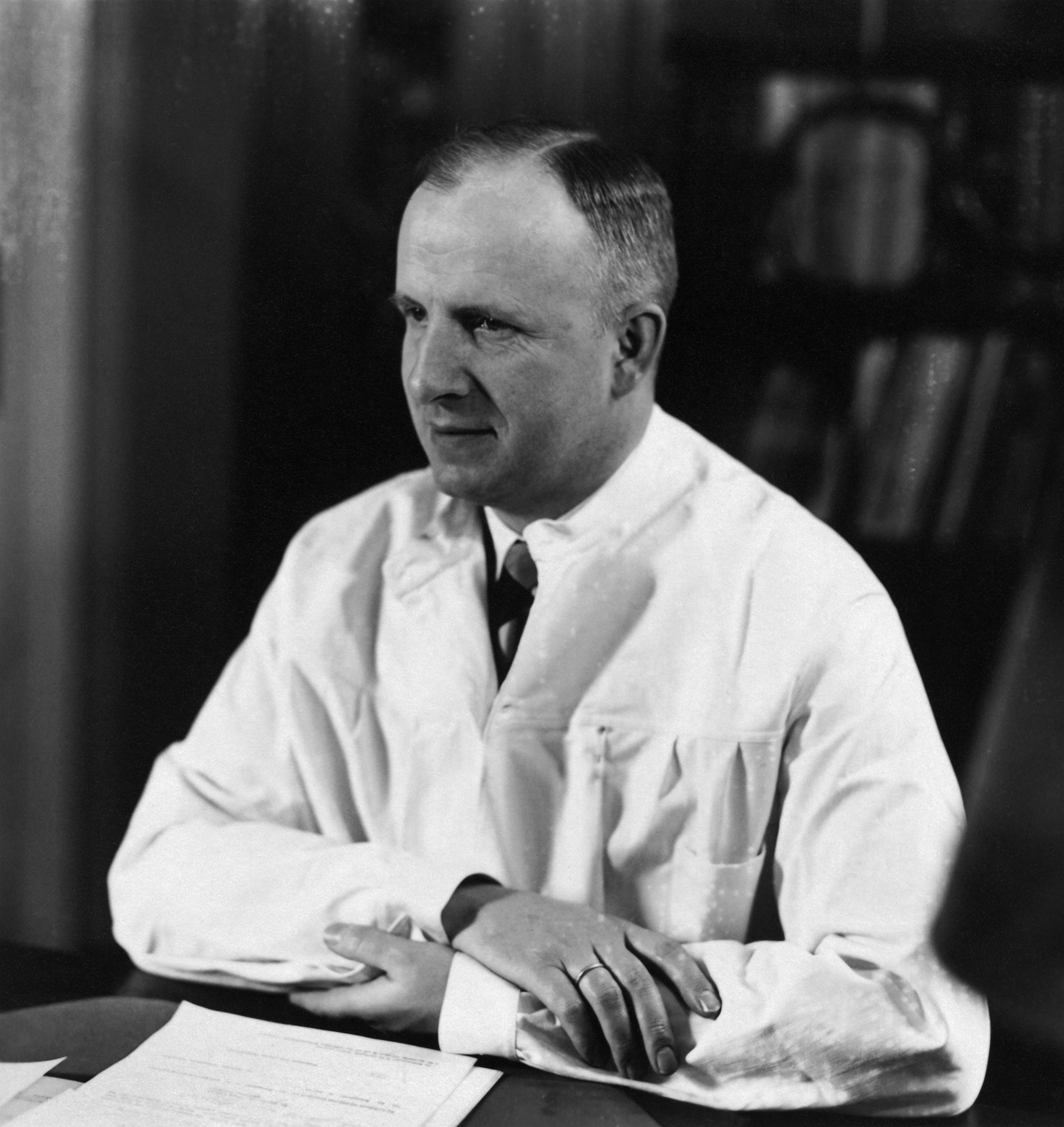 This is a photograph of a White man sitting at a table. His name is Otmar von Verschuer. He is wearing a white lab coat, and his arms are crossed.