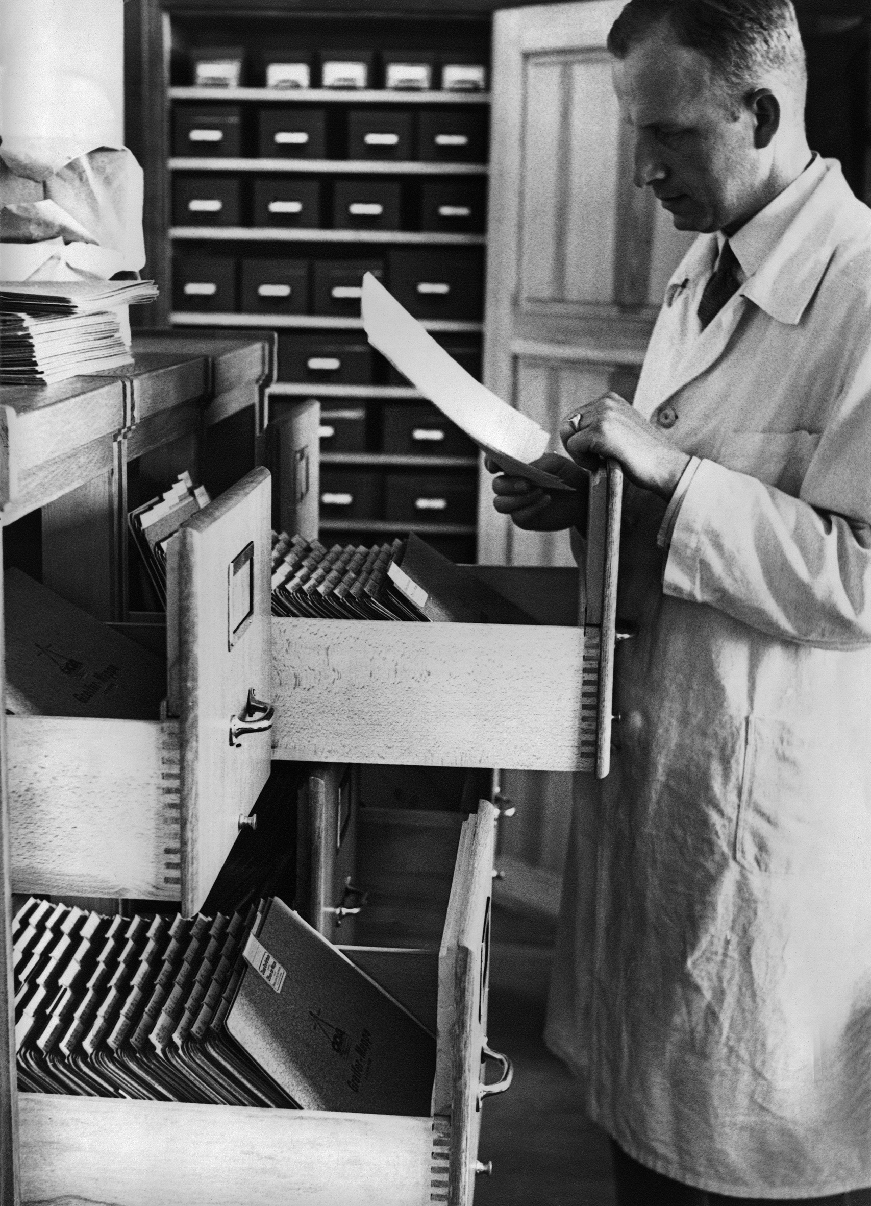 A photograph of a White man wearing a white lab coat. His name is Otmar von Verschuer. He is standing next to an open drawer of a filing cabinet and reading a document from the file on twins.