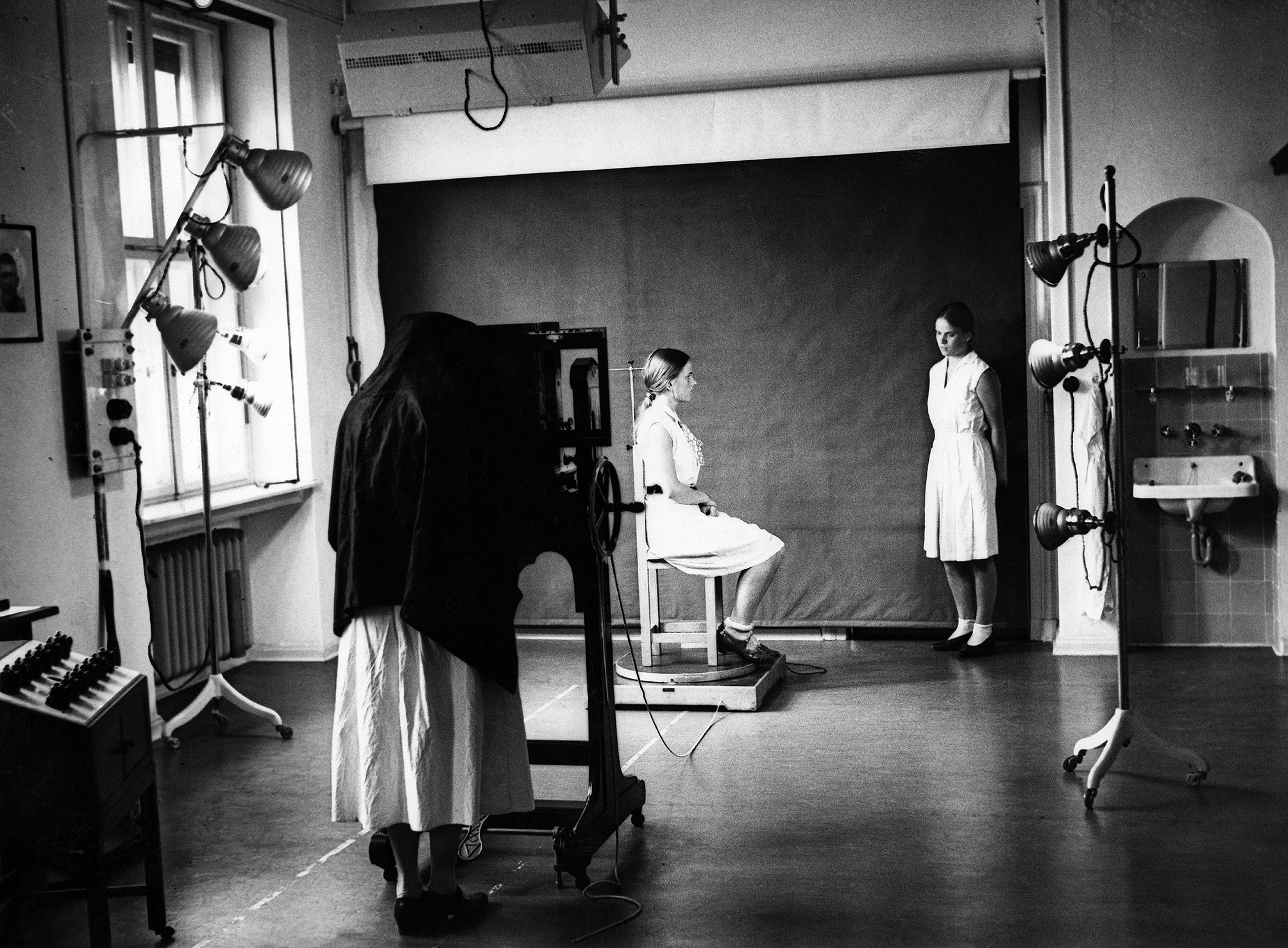 Photograph of three individuals in an examination room. One person wearing a dress or a long white lab coat is taking a picture from the side of a person sitting on a chair. The twin sister of the person being photographed is standing to one side.