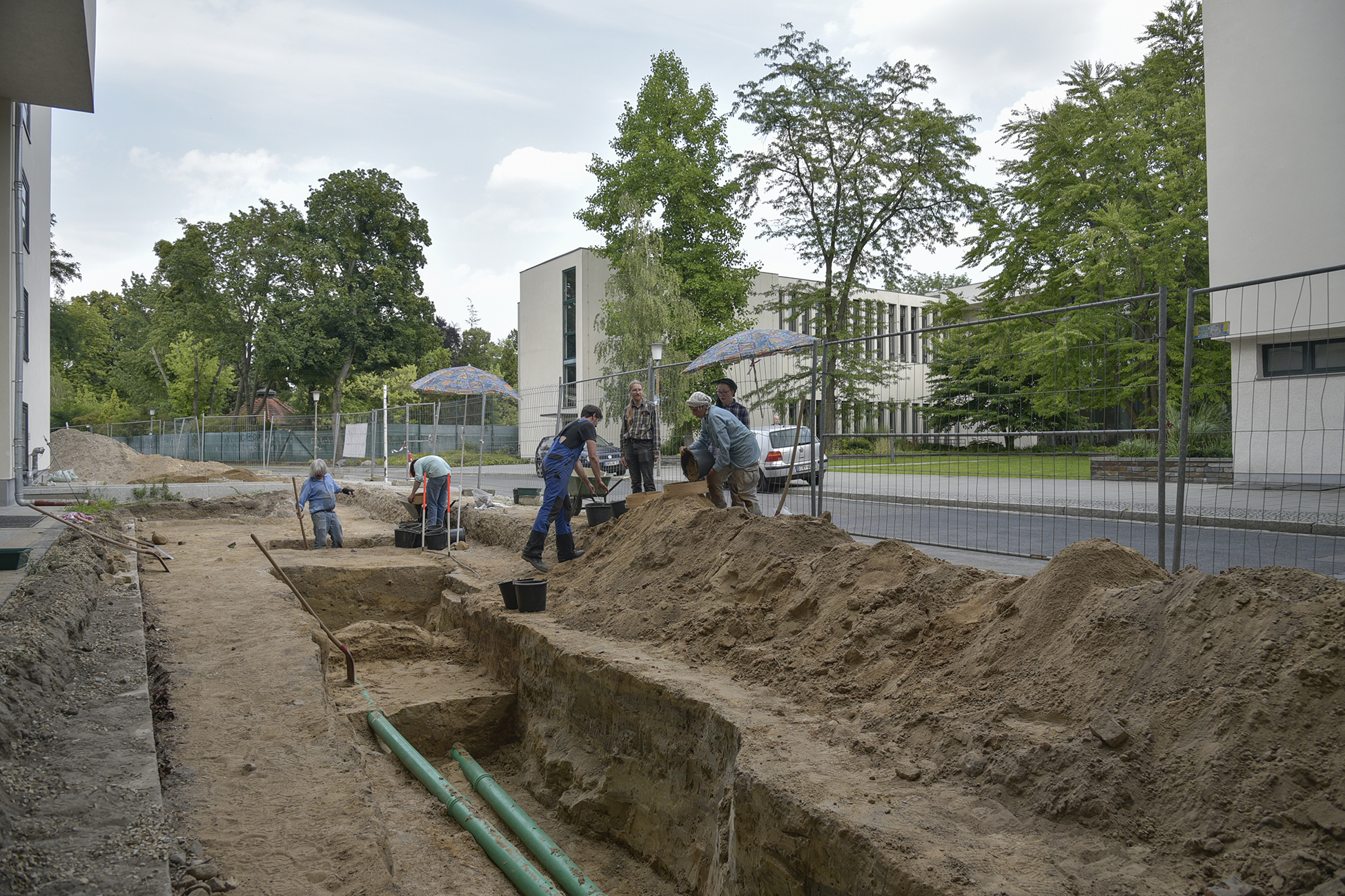 Foto einer archäologischen Grabung. Zwischen einem Bauzaun und einer Hauswand ist ein etwa zwei Meter tiefer und breiter Graben ausgehoben. Sechs Personen in Arbeitskleidung stehen mit Schaufeln und Eimern am Rand des Grabens.