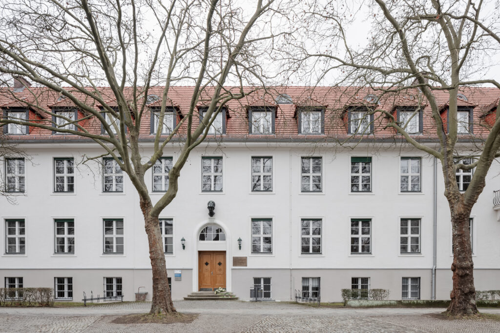 Foto, dass die Vorderansicht des Gebäudes in der Ihnestraße zeigt, Jahr 2024. Über dem runden Torbogen, der den Eingang rahmt, hängt eine metallene Büste der Minerva. Das Haus ist vier Stockwerke hoch, weiß gestrichen und mit rechteckigen Fenstern durchsetzt.