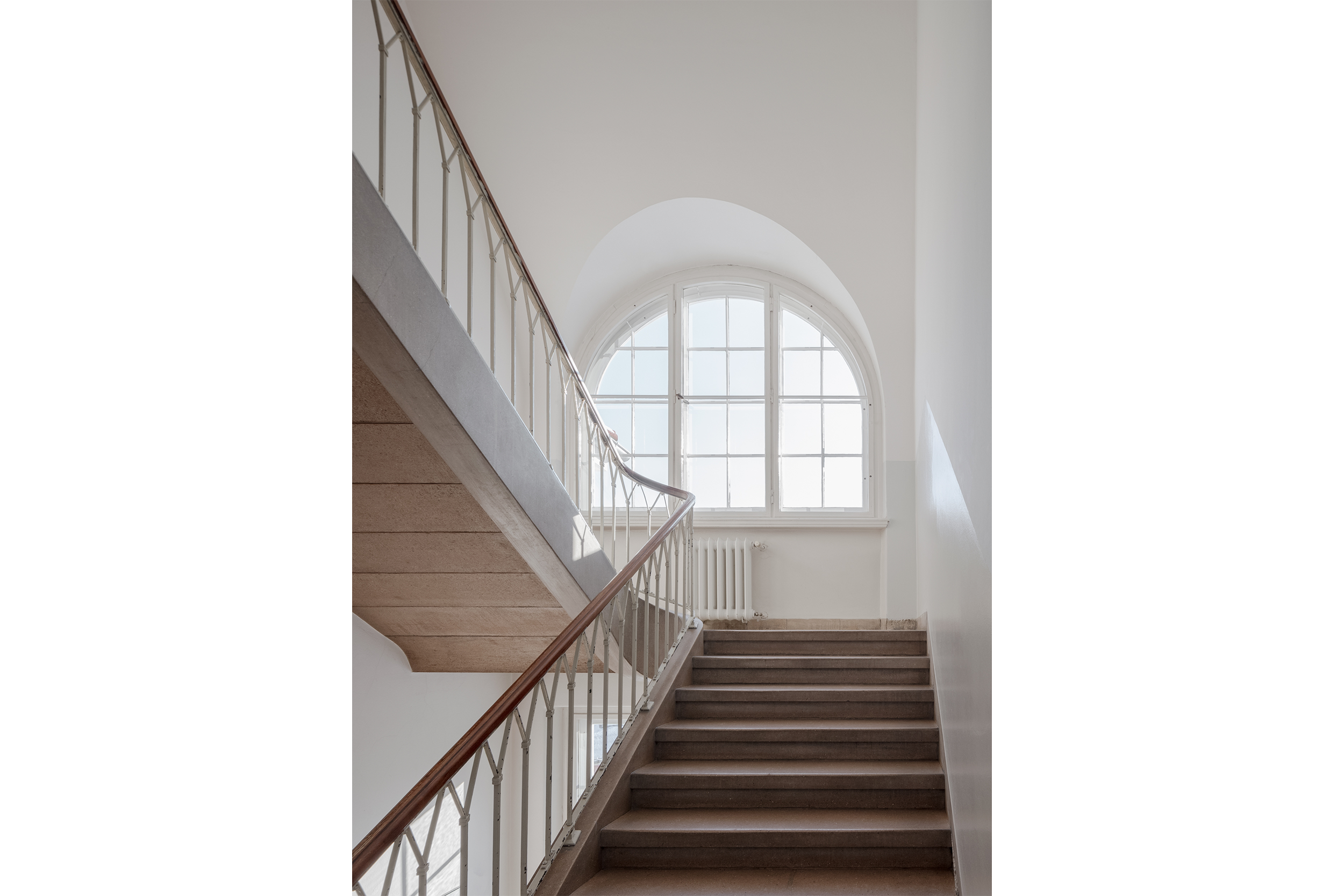 Foto vom Treppenaufgang in der Ihnestraße 22, Jahr 2024. Treppenstufen aus Stein führen zu einer Zwischenetage. Durch ein nach oben hin abgerundetes Fenster fällt Licht auf die Stufen und das Geländer.