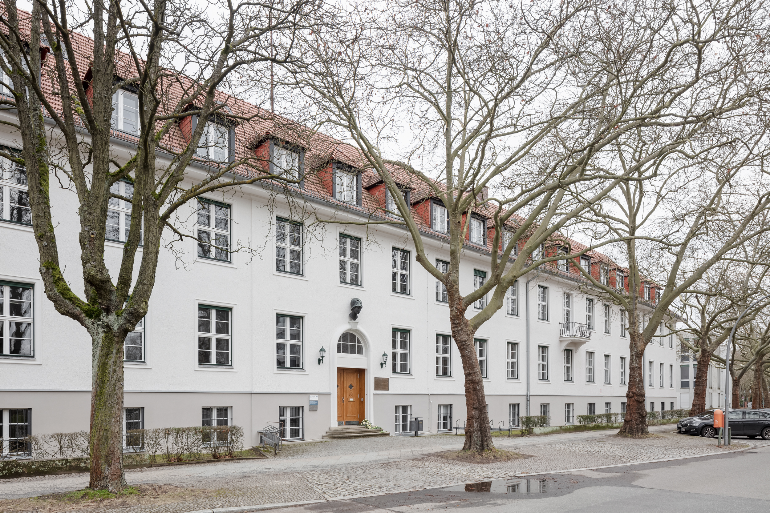 Foto, dass die Vorderansicht des Gebäudes in der Ihnestraße zeigt, Jahr 2024. Über dem runden Torbogen, der den Eingang rahmt, hängt eine metallene Büste der Minerva. Das Haus ist vier Stockwerke hoch, weiß gestrichen und mit rechteckigen Fenstern durchsetzt.